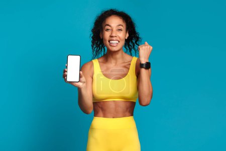 Photo for Workout Applications And Gadgets. Happy black sportswoman showing smartphone with blank screen and fitness smartwatch on wrist, posing over blue studio background, in yellow sportswear. Mockup - Royalty Free Image