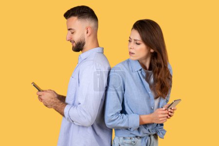 Photo for Woman looks over with curiosity at mans smartphone, while both are engrossed in their devices, creating scene of modern communication on yellow background - Royalty Free Image