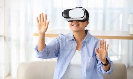 Photo for Joyful asian young woman wearing modern VR glasses while sitting on couch in living room, touching blank space, chinese lady experiencing virtual reality session at home - Royalty Free Image