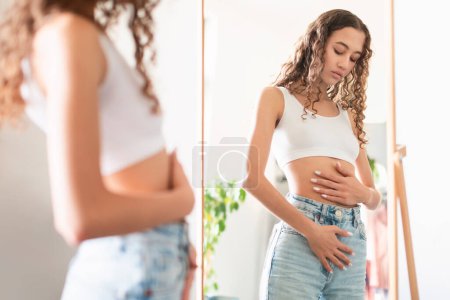 Teenager Girl Touching Her Stomach Suffering From Menstrual Period Pain, Or Showcasing Early Pregnancy Concept, Standing Near Mirror At Home. Abdominal Health. Selective Focus