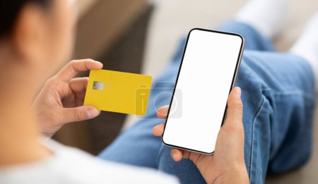 Photo for Over shoulder view of woman using phone with white blank screen mockup copy space and yellow plastic credit card, customer banking or shopping online, paying for services and goods - Royalty Free Image