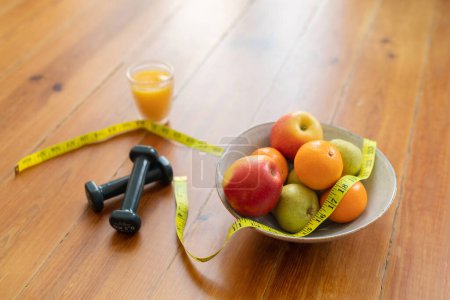 Foto de Dieta y estilo de vida saludable. Cuenco de frutas frescas rodeado por cinta métrica, con pesas y jugo de naranja tumbado en el suelo de madera que simboliza la dieta equilibrada y la rutina de ejercicios, espacio para copiar - Imagen libre de derechos