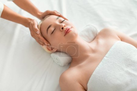 Foto de Facebuilding and Rejuvenation. Mujer rubia europea con los ojos cerrados recibiendo tratamiento de cuidado de la piel y masaje facial en el centro de spa interior, acostado envuelto en toalla blanca - Imagen libre de derechos