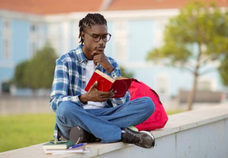 Photo for Focused black young man studying reading book, sitting by the university campus, deep in thought and learning, posing with eyewear and backpack while preparing homework outside - Royalty Free Image