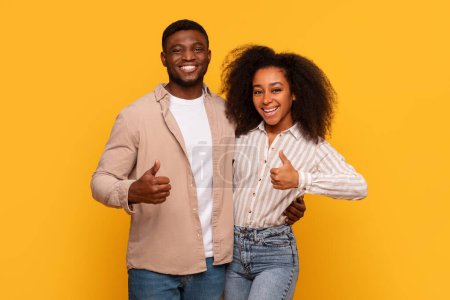Téléchargez les photos : Couple afro-américain souriant montrant l'approbation avec les pouces levés, exsudant des vibrations positives et de la convivialité, dans un contexte jaune joyeux - en image libre de droit
