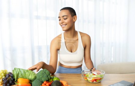 Smiling young latin woman in sportswear with healthy food, salad and vegetables at kitchen interior. Wellness, nutrition, cooking and preparation fresh food at light home