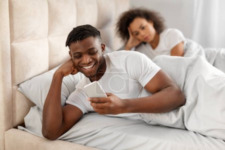 Téléchargez les photos : Phubbing. Femme afro-américaine mécontente regardant son petit ami texter au téléphone au lit, l'ignorant dans la chambre à coucher à la maison. Dame jalouse suspectant une affaire. Focus sélectif - en image libre de droit