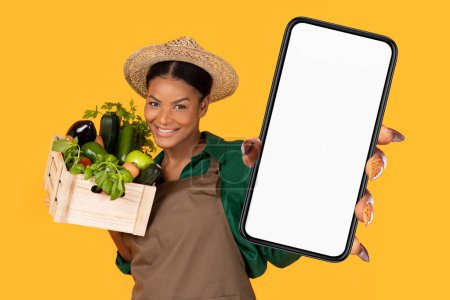Téléchargez les photos : Heureuse dame fermière noire dans le chapeau et tablier tenant la boîte avec la récolte fraîche de sa serre, montrant Big Phone avec écran vide sur fond jaune, la vente de produits en ligne via l'application mobile - en image libre de droit