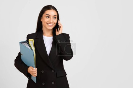 Téléchargez les photos : Femme professionnelle en costume d'affaires parlant au téléphone et tenant des dossiers, regardant loin de l'espace de copie avec le sourire sur un fond propre, occupé au travail - en image libre de droit