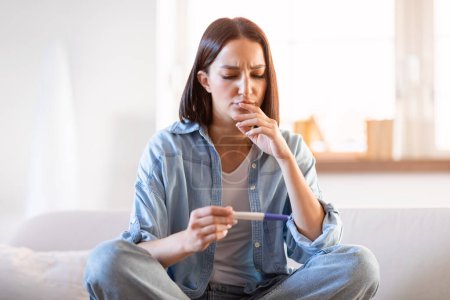 Photo for Unhappy young woman with pregnancy test sitting on couch at home, sad about negative result and infertility or unplanned pregnancies. Problems with conceiving child, female health, unwanted baby - Royalty Free Image