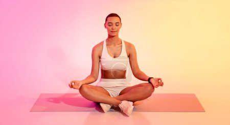 Foto de Mujer joven latina serena con un corte de pelo corto en ropa deportiva meditando en posición de loto sobre una esterilla de yoga, ojos cerrados, sobre un fondo de degradado rosa y amarillo pacífico - Imagen libre de derechos