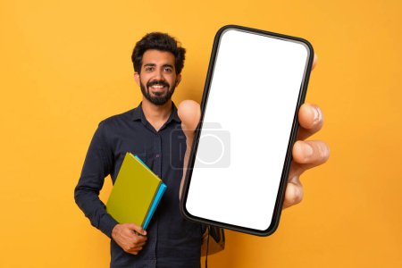 Photo for Online Education, Mobile App. Smiling Young Indian Guy Posing With Workbooks Over Yellow Studio Background, Happy Student Wearing Backpack Showing Phone With White Blank Screen, Copy Space, Mockup - Royalty Free Image