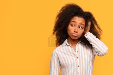 Téléchargez les photos : Jeune femme noire contemplative aux cheveux bouclés regardant loin l'espace libre avec la main sur la tête, montrant une expression de préoccupation ou une pensée profonde sur fond jaune - en image libre de droit