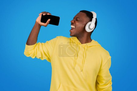 Photo for An African man in a yellow hoodie joyously sings into a smartphone as a microphone, wearing white headphones, against a vibrant blue background, embodying a moment of musical bliss - Royalty Free Image