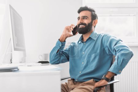 Foto de Relajado y sonriente empresario indio disfrutando de una agradable conversación en su teléfono mientras se inclina de nuevo en su silla de oficina, exudando positividad - Imagen libre de derechos