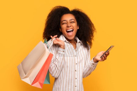 Téléchargez les photos : Délicieuse jeune femme noire aux cheveux bouclés tenant des sacs à provisions colorés et smartphone, riant joyeusement sur fond jaune vif - en image libre de droit