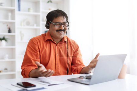Photo for Smiling mature indian man with moustache wearing eyeglasses using laptop and headset, sitting at desk at cozy home office, teach have video call with students. Employee working online, copy space - Royalty Free Image