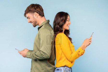 Téléchargez les photos : Conjoints debout dos à dos, chacun absorbé dans leur propre smartphone, symbolisant la connectivité moderne et les mondes numériques individuels, sur fond bleu doux - en image libre de droit