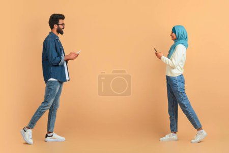 Téléchargez les photos : Couple musulman absorbé dans leurs téléphones portables tout en marchant devant l'autre, jeune homme arabe et femme en hijab debout sur fond beige doux, illustrant la communication moderne, espace de copie - en image libre de droit