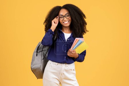 Photo for Cheerful black lady with curly hair, carrying backpack and copybooks, touches her eyeglasses and smiling, posing against yellow background, epitomizing joyful student life - Royalty Free Image