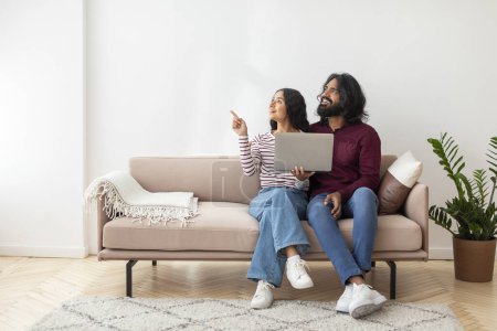 Photo for Positive cheerful millennial indian lovers sitting on couch and using laptop at home, pointing at blank copy space over white wall background, showing nice website, online offer, deal - Royalty Free Image