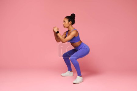 Téléchargez les photos : Concept d'entraînement. Jeune femme noire sportive déterminée faisant un exercice de squat profond pendant l'entraînement sur fond de studio rose. Mode de vie sain, sport et bien-être. Vue latérale - en image libre de droit