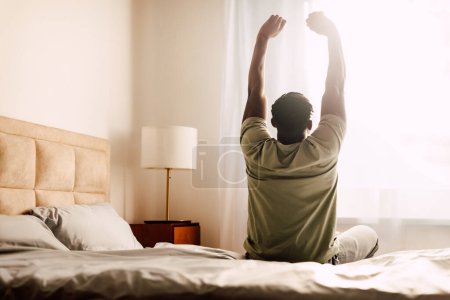 Guten Morgen. Rückenansicht des aufwachenden schwarzen Mannes mit ausgestreckten Armen, der im Bett in einem modernen Schlafzimmer im Haus sitzt und zum Fenster blickt. Beginn des neuen Tages, Wellness und Erholung. Kopierraum