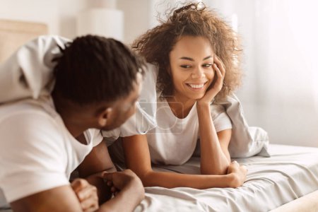 Liebe und Glück. Liebevolle schwarze Ehepartner lächeln einander an und genießen den romantischen Morgen im Schlafzimmer, liegen unter der Decke und flirten mit intimen Momenten im Bett zu Hause