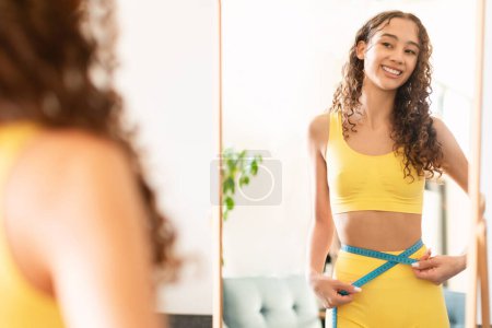 Photo for Smiling caucasian teen girl in sportswear measuring waist with tape, checking inches lost around her belly, feeling proud and happy with her weight loss results, looking at mirror at home - Royalty Free Image