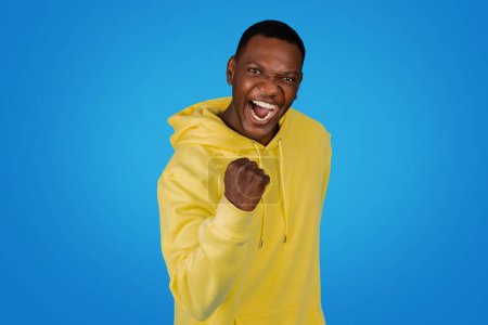 Foto de Un exuberante hombre afroamericano ganador en una sudadera amarilla golpea el aire con un puño cerrado, mostrando una amplia sonrisa victoriosa contra un fondo de estudio azul vivo - Imagen libre de derechos