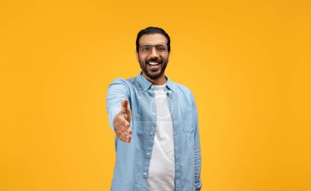 Photo for Amiable man in a denim shirt reaching out for a handshake, smiling warmly, inviting connection and interaction, set against a cheerful yellow background. Deal gesture - Royalty Free Image