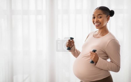 Téléchargez les photos : Femme noire enceinte souriante faisant de l'exercice avec des cloches, regardant la caméra et souriant, panorama avec espace de copie. Les Afro-Américains attendent une formation de dame à la maison, utilisant des haltères. Sport pendant la grossesse - en image libre de droit