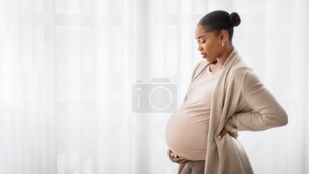 Téléchargez les photos : Joyeux jeune femme noire enceinte en tenue confortable debout à côté de la fenêtre à la maison, regardant son gros ventre, attendant bébé, panorama avec espace de copie. Grossesse, concept de maternité - en image libre de droit