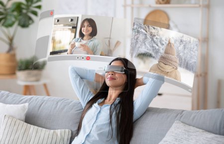 Foto de El futuro de la comunicación está aquí: la joven se entrega a videoconversaciones realzadas por la realidad virtual en casa, relajándose en el sofá con pantallas digitales - Imagen libre de derechos