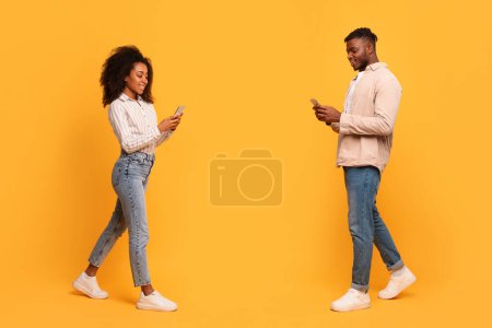 Photo for Smiling young black man and woman walking engrossed in texting on their smartphones against vibrant yellow backdrop, full length - Royalty Free Image