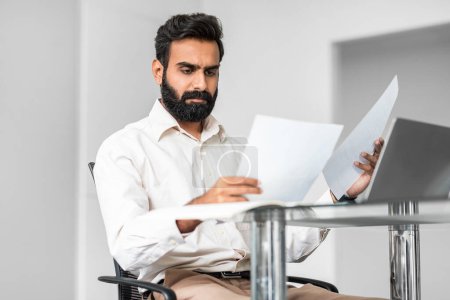 Photo for Serious businessman in a white shirt, meticulously reviewing printed reports while sitting at a glass table in a modern office, displaying concentration and attention to detail - Royalty Free Image