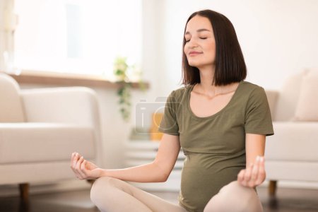 Foto de La mujer embarazada se sienta en posición de loto en una esterilla de yoga en el interior de casa, sus ojos cerrados en meditación tranquila, centrándose en la respiración profunda para el bienestar del feto. Embarazo y relajación - Imagen libre de derechos