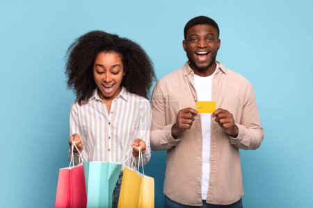 Joyeux couple afro-américain avec plusieurs sacs à provisions et carte de crédit en or, célébrant la journée amusante de la thérapie au détail, sur fond bleu vibrant, exsudant bonheur et joie des consommateurs