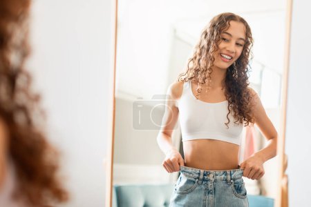 Téléchargez les photos : Jeune adolescente heureuse vérifiant ses progrès minceur, tirant la taille de son gros jean, se tenant près du miroir, célébrant la perte de poids réussie. Concept de réussite de l'alimentation - en image libre de droit