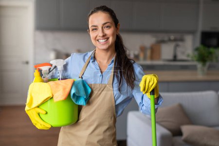 Foto de Mujer feliz equipada con fregona y cubo lleno de suministros de limpieza destellos sonrisa cálida, listo para hacer frente a las tareas del hogar con confianza - Imagen libre de derechos
