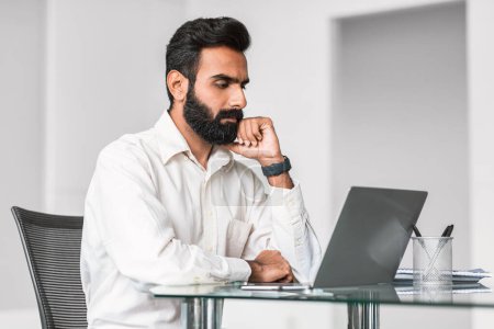 Besinnlicher indischer Profi mit Bart, der an einem gläsernen Schreibtisch über die Arbeit auf seinem Laptop sinniert und fokussierte Problemlösung in einem modernen Büro verkörpert