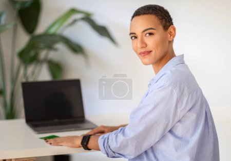 Foto de Una joven latina profesional sonriente con el pelo corto posa con confianza junto a un escritorio con una computadora portátil, encarnando la elegancia y eficiencia del lugar de trabajo moderno, disfruta del trabajo, el estudio - Imagen libre de derechos