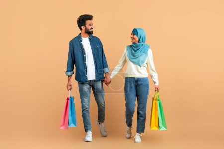 Téléchargez les photos : Couple musulman appréciant faire du shopping ensemble, se tenant la main et portant des sacs colorés, conjoints arabes avec des sourires contentés marchant sur fond studio beige, symbolisant la joie des consommateurs - en image libre de droit