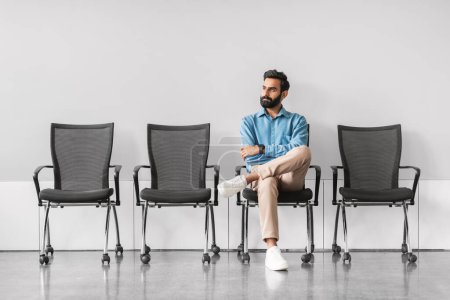 Photo for Composed indian businessman seated with legs crossed, reflecting alone in an empty row of chairs in a sparse and modern waiting area, full length - Royalty Free Image