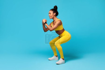 Photo for Fitness And Sport. Smiling Slim Black Young Woman With Earbuds And Smartwatch Doing Deep Squat, Exercising During Workout, Warming Up Over Blue Studio Background. Full Length - Royalty Free Image