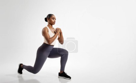 Afroamerikanische Fitness-Frau streckt die Beine nach vorne und springt über die weiße Studiokulisse, genießt die Workout-Routine, schaut zur Seite. Ganzer Schuss, leerer Raum