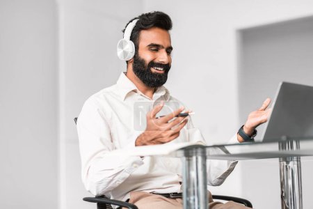 Téléchargez les photos : Délicieux homme d'affaires indien en chemise blanche utilisant des écouteurs pour un appel vidéo animé sur son smartphone, exprimant sa positivité dans une configuration de bureau moderne - en image libre de droit