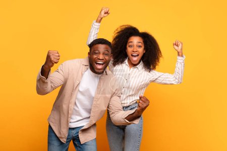 Photo for Overjoyed black man and woman with raised fists, wide smiles, and eyes shining with excitement, celebrating joyful moment together against cheerful yellow backdrop - Royalty Free Image