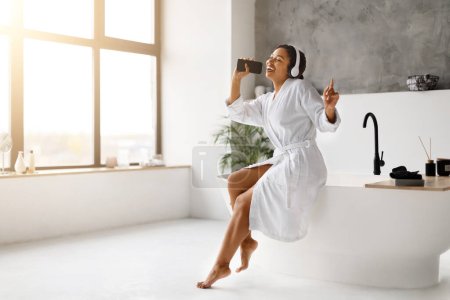 Femme noire en peignoir blanc chantant dans son téléphone comme microphone, portant des écouteurs, femme afro-américaine joyeuse assise sur la baignoire, relaxant dans l'intérieur de la salle de bain ensoleillée, espace de copie