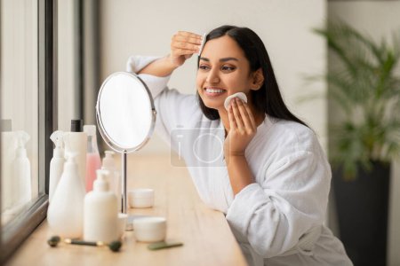 Téléchargez les photos : Belle femme indienne souriante nettoyer la peau avec des tampons de coton tout en faisant routine quotidienne de beauté dans la salle de bain, regardant vers le miroir, attrayant jeune femme bénéficiant de soins de la peau à la maison, Copier l'espace - en image libre de droit